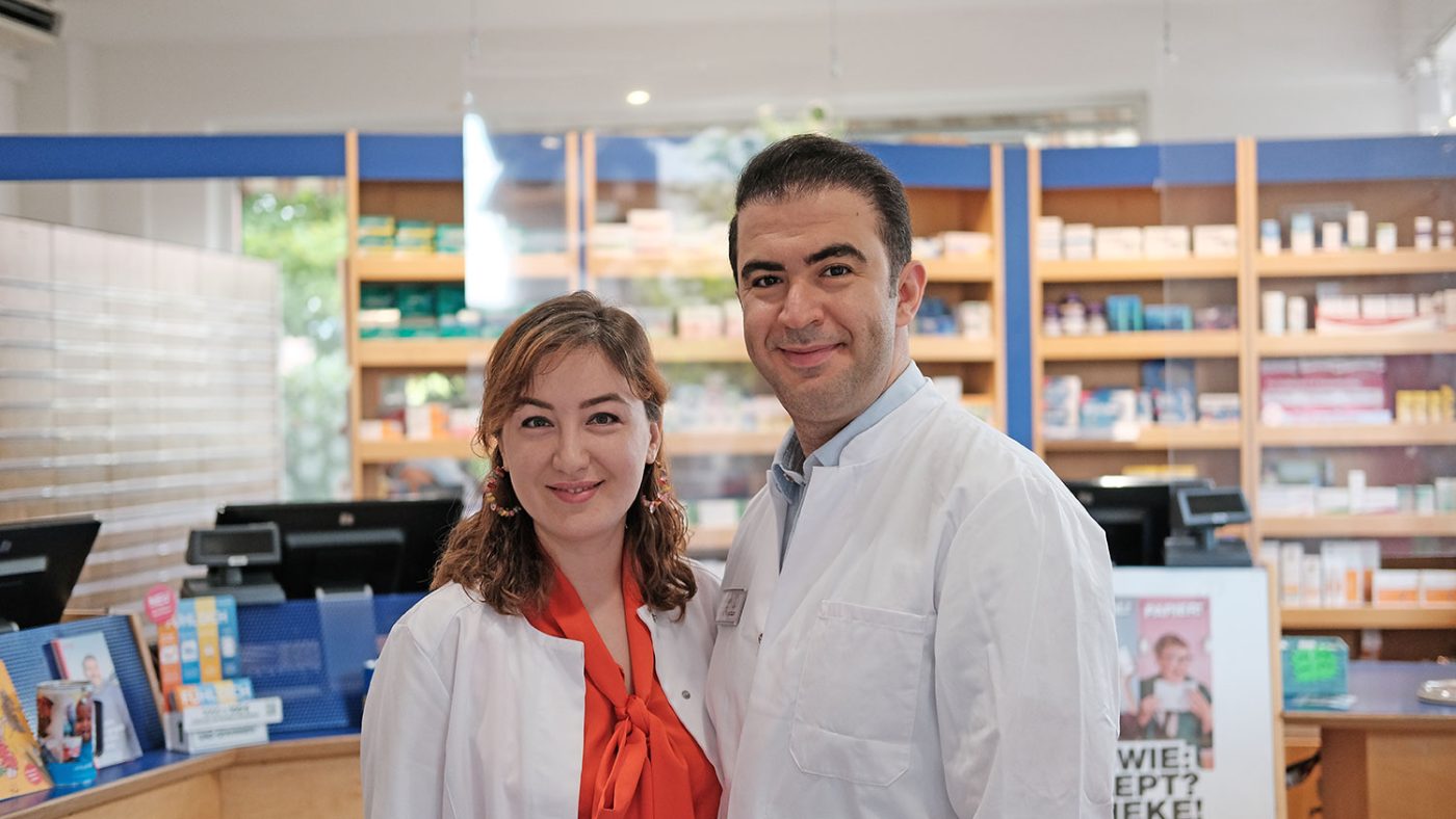Apothekerin Saba Maleki-Nejatian und Dr. Nojan Nejatian in weißen Kitteln vor den Regalen im Verkaufsraum der Heegbach Apotheke