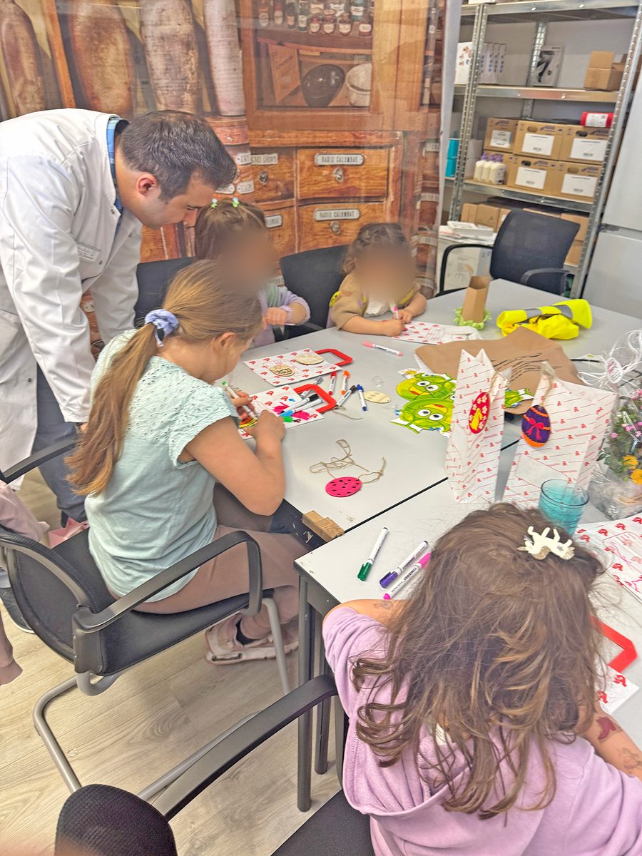 Kinder malen und basteln am Tisch im Kids Club der Heegbach Apotheke, ein Apotheker im weißen Kittel hilft ihnen